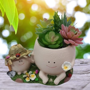 May include: A white ceramic planter shaped like a smiling person with legs and arms, holding a small white flower. The planter is filled with a variety of green succulents. A smaller ceramic planter shaped like a smiling person with a straw hat sits next to the larger planter.