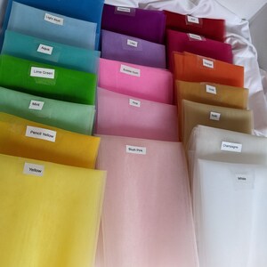 May include: A collection of 16 different colored tulle fabrics, each with a label indicating its color. The colors include royal blue, turquoise, light blue, aqua, lime green, mint, pencil yellow, yellow, magenta, lilac, bubble gum, pink, gold, nude, blush pink, champagne, and white.
