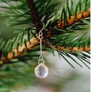 Puede incluir: Un solo pendiente de plata con un colgante de cristal transparente facetado. El pendiente cuelga de un gancho y una cadena de plata, sobre un fondo de agujas de pino verdes y ramas marrones.