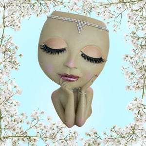 May include: A beige-colored face-shaped planter with closed eyes, long eyelashes, and a jeweled headband. The planter has a pink blush and a red lip color. The hands are placed under the chin, and the planter is surrounded by white flowers.