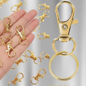 May include: Gold-colored metal swivel clasps and key rings. The clasps have a lobster claw design and a small ring for attaching items. The key rings are round and shiny. Several clasps are scattered around a hand.