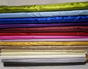 Op de afbeelding: Een stapel van 12 verschillende gekleurde satijnen stoffen. De kleuren zijn groen, lichtblauw, blauw, paars, bordeauxrood, roze, goud, beige, crème, wit, grijs en zwart.
