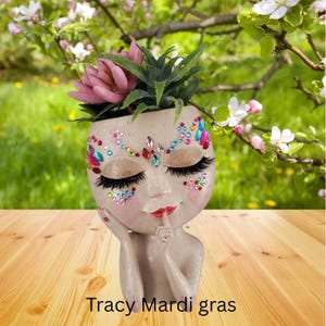 May include: A white ceramic planter shaped like a woman's face with closed eyes and a pink flower on top. The face is decorated with colorful rhinestones and the planter has a finger to its lips. The text "Tracy Mardi gras" is written below the planter.