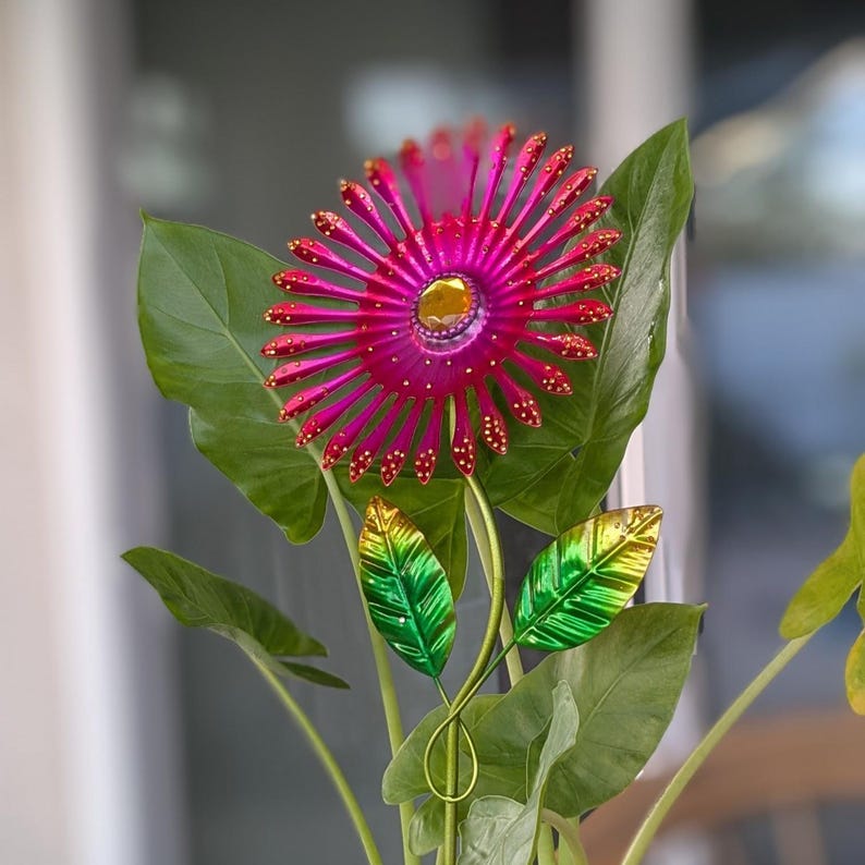 May include: A decorative metal flower with a magenta centre and radiating petals. The flower has a gold centre and is accented with small red dots. Green and gold leaves and stems complete the floral design, set against large green leaves.