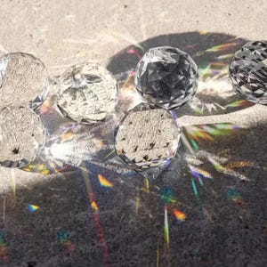 May include: A collection of clear, faceted crystal balls casting colorful light refractions on a textured gray surface. The spheres vary in clarity and surface texture, creating a play of light and shadow.