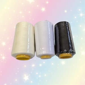 Könnte beinhalten: Drei Garnrollen in Off-White, Weiß und Schwarz, verpackt in Klarsichtfolie. Die Spulen haben gelbe Basen. Der Hintergrund ist ein weicher Farbverlauf aus Rosa, Blau und Gelb mit Sternen.