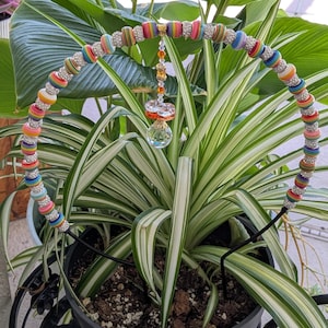 May include: A circular plant trellis featuring a black metal frame adorned with alternating rainbow-striped beads and silver-toned rhinestone-encrusted spacer beads. A dangling ornament with orange and clear faceted crystals hangs from the center.