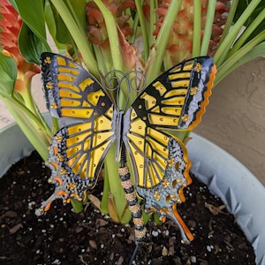 May include: A decorative metal butterfly garden stake with yellow, black, orange, and silver details. The butterfly has a detailed wing pattern and is embellished with small, sparkling accents. The stake is placed in a potted plant, adding a touch of whimsy to the garden.