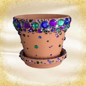 May include: A terracotta plant pot and matching saucer decorated with scattered round rhinestones in shades of blue, green, and purple. The rim of the pot features a dense band of these colorful gems, while the body and saucer have smaller accents.