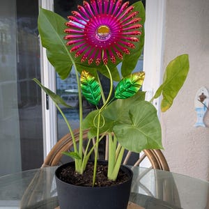 May include: A potted plant with large green leaves and a vibrant, metallic pink and gold flower sculpture. The flower has a central yellow gem and red accents. The pot is black and sits on a wooden base.