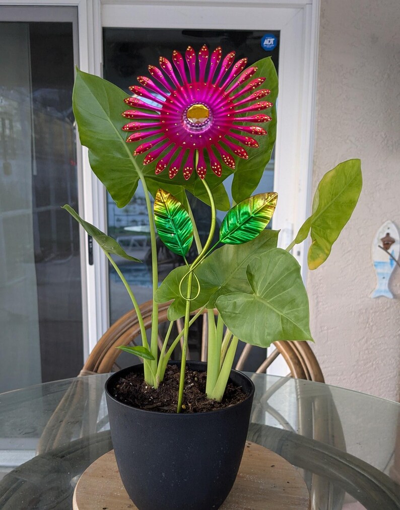 May include: A potted plant with large green leaves and a vibrant, metallic pink and gold flower sculpture. The flower has a central yellow gem and red accents. The pot is black and sits on a wooden base.