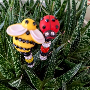May include: Two decorative garden stakes featuring a bee and a ladybug. The bee has a yellow body with black accents and white wings. The ladybug is red with black spots. Both have jeweled stems with alternating colors of black, silver, yellow, and red.