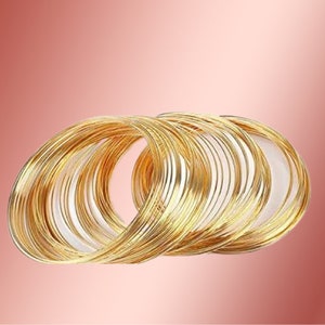 May include: A set of multiple thin, circular gold-colored bangles stacked together. Each bangle features a smooth, polished metallic finish with a reflective surface. The bracelets are arranged in an overlapping cluster, creating a layered appearance.