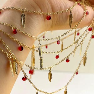 May include: Gold-toned layered necklace featuring delicate chains adorned with small red beads and feather charms. The necklace is designed with multiple strands, creating a layered effect. The feather charms add a touch of bohemian flair to this jewelry piece.
