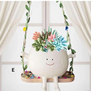 May include: A whimsical white ceramic planter shaped like a smiling face, suspended on a wooden swing. The planter is filled with colorful faux succulents. The swing is decorated with green vines and small flowers.