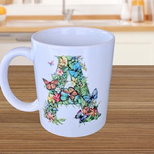 Pode incluir: Caneca de cerâmica branca com um design floral e de borboletas em forma da letra "A". O design inclui borboletas e flores coloridas em tons de rosa, laranja, azul e amarelo. A caneca tem uma alça curva.