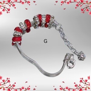 May include: A silver bracelet with red and clear crystal beads. The bracelet has a chain extension and a decorative clasp. The letter "G" is visible on the background.