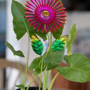 May include: A decorative metal flower sculpture with a magenta and gold centre, surrounded by magenta petals with gold accents. The sculpture has green metal leaves and a slender stem, set in a potted plant with large green leaves.