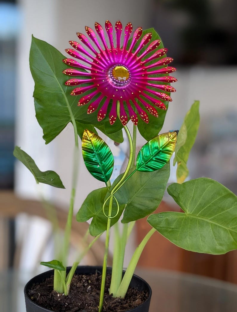 May include: A decorative metal flower sculpture with a magenta and gold centre, surrounded by magenta petals with gold accents. The sculpture has green metal leaves and a slender stem, set in a potted plant with large green leaves.