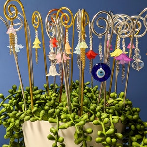 Può includere: Una collezione di paletti da giardino decorativi in metallo con ciondoli di fiori colorati e un ciondolo blu di occhio turco. I paletti sono disposti in un vaso di fogliame verde.