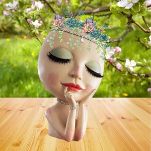 May include: A decorative face planter with closed eyes, red lips, and a floral crown. The planter has a beige complexion with pink blush and is adorned with a rhinestone necklace and bracelet. The background features a wooden surface and a blurred green and floral backdrop.