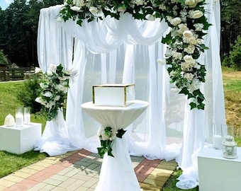 May include: White wedding ceremony backdrop with a floral arch made of white flowers and greenery. The backdrop is made of white sheer fabric and is draped over a metal frame. A white table with a white tablecloth and a white box on top is in front of the backdrop.
