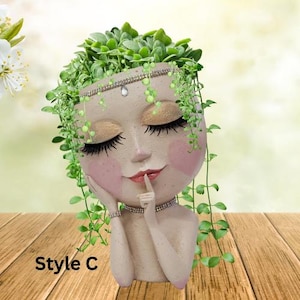 May include: A white ceramic planter shaped like a woman's face with closed eyes and a finger to her lips. The planter has a beaded headband and is filled with green plants.