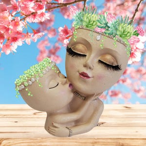 May include: A pair of ceramic planter faces, one with a crown of green succulents, the other with a pearl necklace. The faces are painted with closed eyes, eyelashes, and lips. The larger face is kissing the smaller face on the cheek.