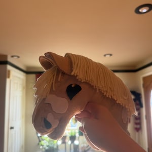 Pode incluir: Uma cabeça de cavalo de pelúcia em tons de bege e creme. O brinquedo apresenta uma crina creme, olhos pretos e marcas brancas no rosto. A cabeça de cavalo é projetada para ser segurada e brincada.