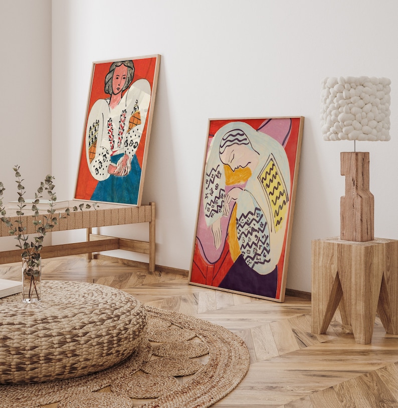 May include: Two framed paintings of women in white shirts with colorful patterns, hanging on a white wall. A white fluffy lamp sits on a wooden stool.