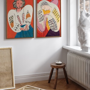 May include: Two framed paintings of women in colorful clothing with geometric patterns. The paintings are hung on a white wall with a wooden floor.