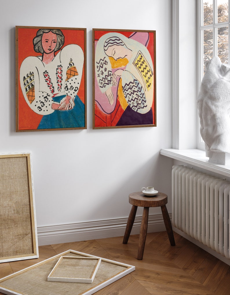 May include: Two framed paintings of women in colorful clothing with geometric patterns. The paintings are hung on a white wall with a wooden floor.