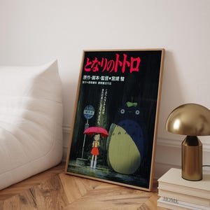 May include: Framed poster featuring the animated film "My Neighbor Totoro." The artwork depicts a young girl with a red umbrella, Totoro, and Japanese text. The poster is in a wooden frame and leans against a white wall.