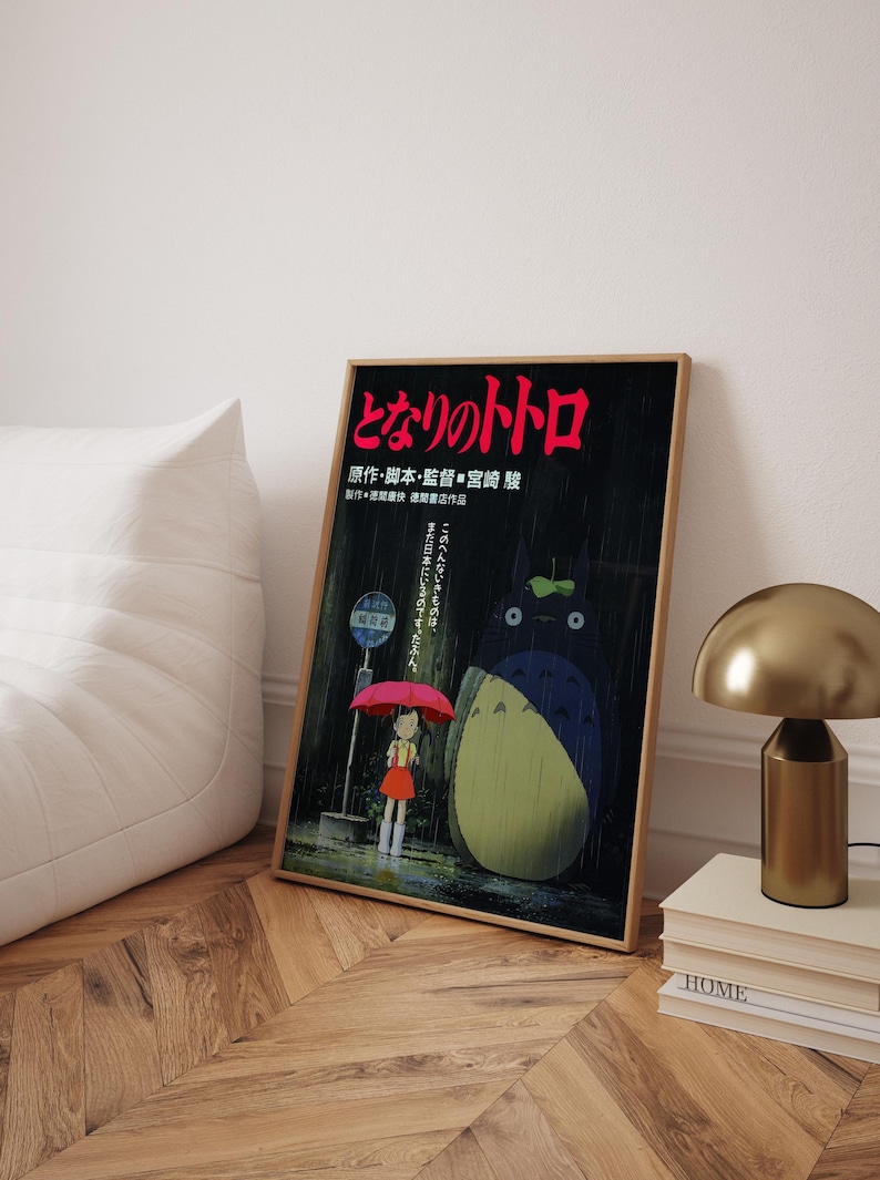May include: Framed poster featuring the animated film "My Neighbor Totoro." The artwork depicts a young girl with a red umbrella, Totoro, and Japanese text. The poster is in a wooden frame and leans against a white wall.