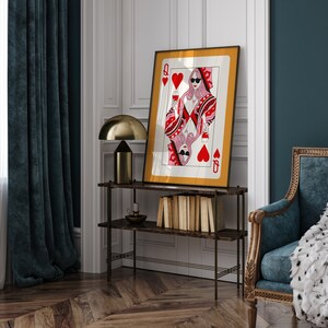 May include: A framed print of a Queen of Hearts playing card with a woman in sunglasses holding a glass of wine. The card is surrounded by a gold frame and the background is a white wall.