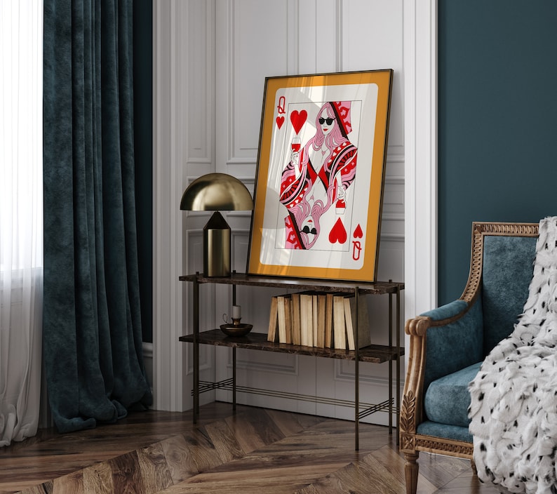 May include: A framed print of a Queen of Hearts playing card with a woman in sunglasses holding a glass of wine. The card is surrounded by a gold frame and the background is a white wall.