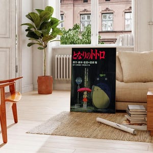 May include: A framed poster featuring the animated film "My Neighbor Totoro" is propped against a beige sofa. The poster shows a girl with an umbrella and Totoro. A potted plant and wooden furniture complete the room.