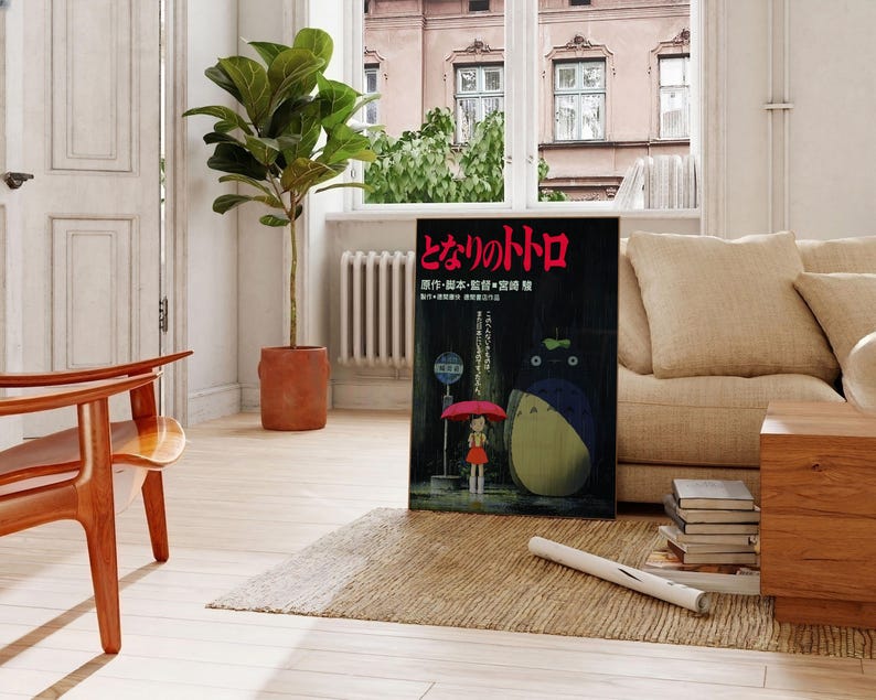 May include: A framed poster featuring the animated film "My Neighbor Totoro" is propped against a beige sofa. The poster shows a girl with an umbrella and Totoro. A potted plant and wooden furniture complete the room.