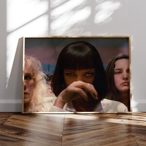 May include: A close-up shot of three women, the woman in the center has dark hair and is looking directly at the camera with a serious expression. The woman on the left has blonde hair and is looking to the right. The woman on the right has dark hair and is looking to the left. The women are all wearing white shirts.