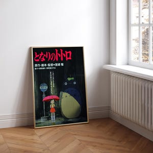 May include: Framed poster featuring the Japanese animated film "My Neighbor Totoro." The artwork depicts a young girl with a red umbrella standing at a bus stop with Totoro. The poster has Japanese text in red and gold tones.
