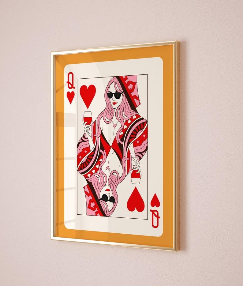 May include: A framed print of a Queen of Hearts playing card with a woman wearing sunglasses and holding a glass of wine. The card is red and pink with a gold frame.