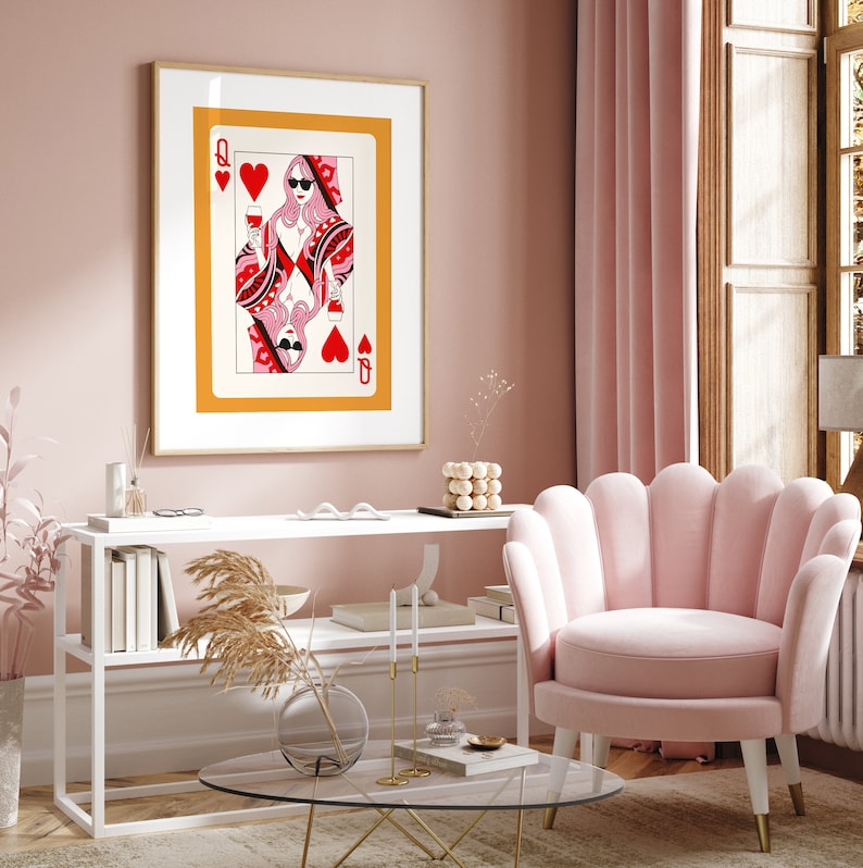 May include: A framed print of a Queen of Hearts playing card with a woman wearing sunglasses and a red dress holding a glass of wine. The print is hanging on a pink wall above a white shelf with books and a vase with dried flowers. A pink armchair sits in front of the shelf.