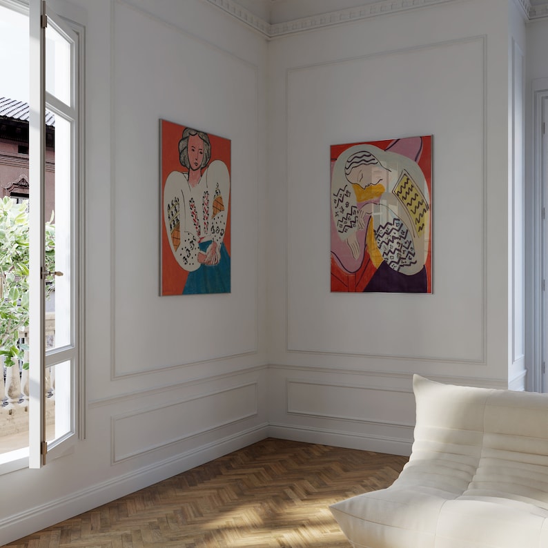 May include: Two framed paintings of women in white shirts with colorful patterns on a white wall. The paintings are in a modern living room with a white couch.