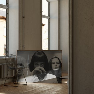 May include: A black and white photograph of three people, two women and one man, in a framed poster. The women are looking at the camera, while the man is looking down. The poster is hanging on a white wall.