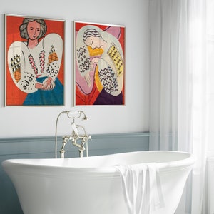 May include: Two framed paintings of women in colorful clothing. The paintings are hanging on a white wall above a white bathtub.