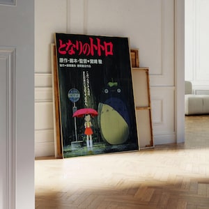 May include: A framed poster featuring Japanese text and a colorful illustration of a girl with a red umbrella and a large, furry creature. The poster is leaning against a white wall and a wooden frame.
