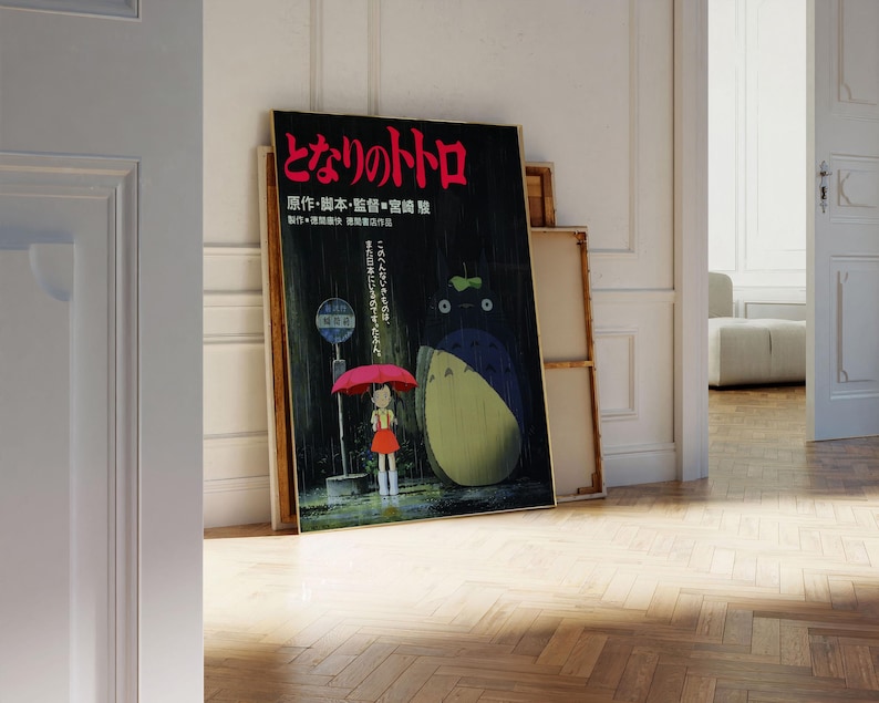 May include: A framed poster featuring Japanese text and a colorful illustration of a girl with a red umbrella and a large, furry creature. The poster is leaning against a white wall and a wooden frame.