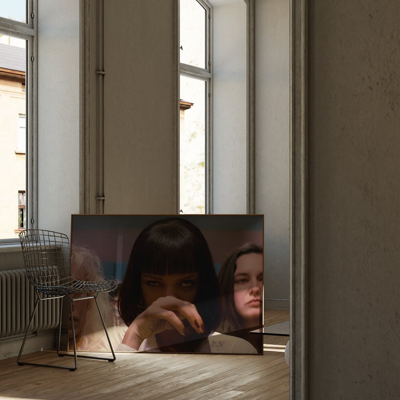 May include: A framed poster featuring a close-up of a woman with dark hair and a serious expression. She is wearing a black shirt and has her hand covering her mouth. A second woman with long dark hair is visible in the background.