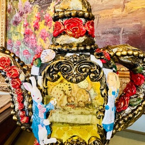 May include: Ornate, gold-painted teapot sculpture featuring red roses, a checkered pattern, and figures from Alice in Wonderland. The teapot has a clock face and a detailed, decorative handle.