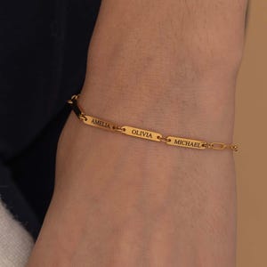 May include: A gold-colored bracelet with the names "AMELIA", "OLIVIA", and "MICHAEL" engraved on it. The bracelet is worn on a wrist, with a white sleeve visible in the background. The bracelet is a delicate piece of jewelry.